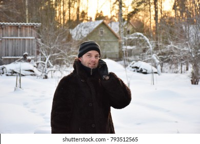 Russian Man In A Winter Coat Old Talking On The Phone In The Village Lit By The Sun At Sunset. Leningrad Oblast, Russia