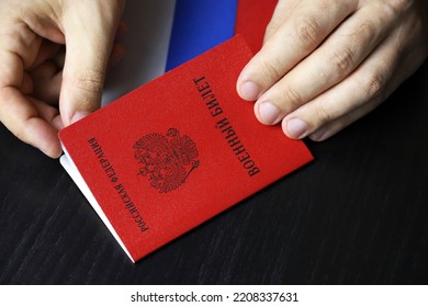 Russian Military ID In Male Hands On Wooden Table. Translation Of Inscription Military ID Card, Russian Federation. Concept Of Mobilization In Russia