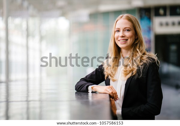 A Russian
woman in a suit leans against a stone in the day and is smiling.
She is blond with blue eyes and is attractive and professional in a
black suit and white shirt. 