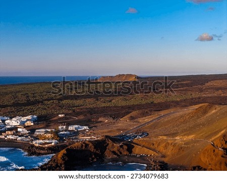 Rust colored volcanic cliffs, black basalt, and ochre cones frame a whitewashed village by a cove near El Golfo and Charco Verde in Lanzarote, with walkers on a road.