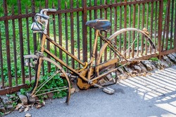 A Rusted, Abandoned Bicycle Leans Against A Metal Fence, Its Journey Halted. The Wheels Are Missing, And Nature Begins To Reclaim The Space Around It, Symbolizing Neglect And The Passage Of Time.