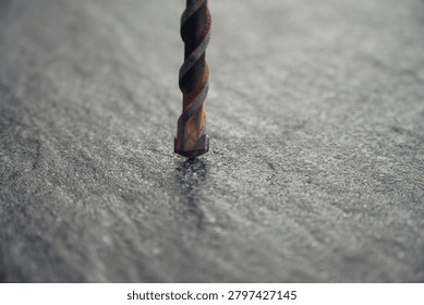 Rusted drill bit stands vertically on a rough surface. Gray, textured background highlights the tool's worn state. Close-up view shows detailed metal spiral and rust. Ideal for construction, repair