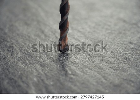 Rusted drill bit stands vertically on a rough surface. Gray, textured background highlights the tool's worn state. Close-up view shows detailed metal spiral and rust. Ideal for construction, repair