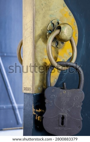 Rusted lock hangs on aged, blue-painted wooden door. Vintage ring and padlock show signs of wear and time. Muted tones suggest abandonment or quiet history. Ideal for themes of closure, memory