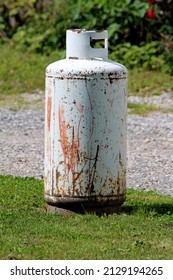 Rusted Old Light Grey Propane Tank Or Grill Cylinder Used For Powering Barbeque Grill Left Outside Of Suburban Family House On Freshly Cut Grass Next To Gravel Driveway