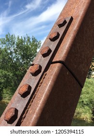 Rusted Rivets And Bolts On A Steel Girder Of Foot Bridge With Rusty Brown Coloring, Metal Connector Plates, Shadows And Sky And Trees In Background.