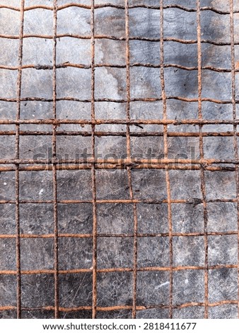Rusted wire mesh grid with a square pattern. The deep orange-brown rust covers the metal against a worn, dark grey, scratched background. Small particles are trapped in the mesh.