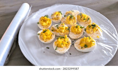 Rustic 1970s style deviled eggs covered in plastic wrap, ready to store in the fridge or take to a picnic. Close-up of homemade curried eggs sealed with clingfilm to keep fresh for a traditional outdoor party or buffet. Isolated image of fresh organic, free-range eggs with bright yellow yolks made in to curried or devilled egg-halves, seasoned and garnished. Rustic, authentic food photograph of  no piping bag or piping skills required, easy hand blended filling for traditional, basic, 1970s vibe appetizer or side salad dish. Authentic, imperfect curried eggs covered with plastic wrap or clingfilm to store in fridge for later, or to put outside on a picnic table. Wrapped or wrapping food in clingfilm or plastic wrap for refrigerator storage.