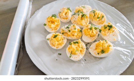 Rustic 1970s style deviled eggs covered in plastic wrap, ready to store in the fridge or take to a picnic. Close-up of homemade curried eggs sealed with clingfilm to keep fresh for a traditional outdoor party or buffet. Isolated image of fresh organic, free-range eggs with bright yellow yolks made in to curried or devilled egg-halves, seasoned and garnished. Rustic, authentic food photograph of  no piping bag or piping skills required, easy hand blended filling for traditional, basic, 1970s vibe appetizer or side salad dish. Authentic, imperfect curried eggs covered with plastic wrap or clingfilm to store in fridge for later, or to put outside on a picnic table. Wrapped or wrapping food in clingfilm or plastic wrap for refrigerator storage.
