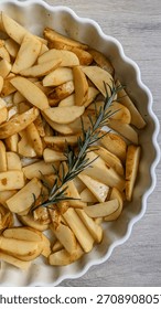 A rustic and appetizing preparation of raw potato wedges placed in a white scalloped baking dish, seasoned lightly and garnished with sprigs of fresh rosemary. 