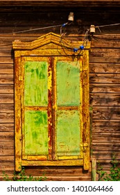 Rustic architectural elegance: weathered cabin wall showcases vintage hardwood window with character, revealing passage of time through distressed surfaces. Vibrant yet muted green shutters display peeling edges that expose rustic yellow undertones, adding textural depth and historic charm. Intricate decorative pediment crowns the window, enhancing quaint old-world aesthetic with ornamental craftsmanship. Horizontal lumber planks in natural brown tones create siding that complements the scene, establishing warm inviting atmosphere. Electrical wires suspend small hanging light fixtures overhead, suggesting practical vintage living while dappled greenery backdrop hints at nature's harmonious encroachment. Soft natural illumination casts gentle shadows, highlighting wood grain and paint imperfections that evoke nostalgic mood. Central framing with ample negative space accommodates text overlays for marketing concepts.