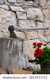 Rustic axe embedded in a weathered chopping block beside a stone wall, with bright red geranium flowers adding color to this traditional outdoor still life.