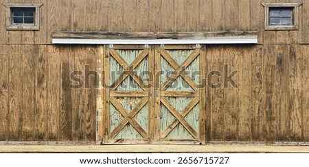 A rustic barn door with a double sliding mechanism, featuring weathered wood, diagonal cross-bracing, and two small windows above. The aged texture adds farmhouse charm.
