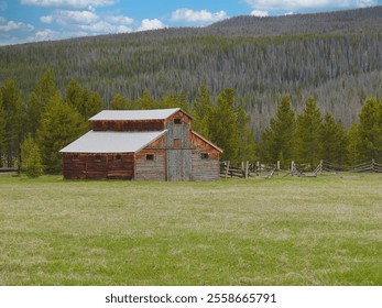 A rustic barn stands in the middle of a lush green field, surrounded by dense pine forests and mountains in the background. The sky is partly cloudy, casting soft shadows on the landscape. - Powered by Shutterstock