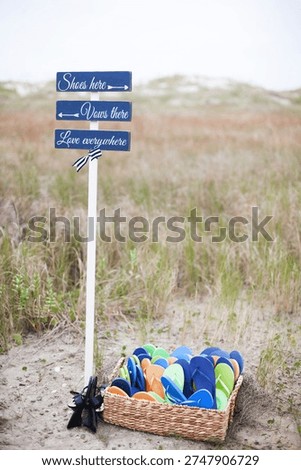 Rustic beach wedding sign reading “Shoes here, vows there, love everywhere” with arrow, wooden directional sign on sandy path with dune grass, romantic coastal ceremony decor.