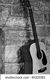 Rustic black and white image of a steel-stringed acoustic guitar leaning against a stone wall.