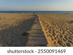 A rustic boardwalk stretches across the sandy beach, inviting visitors toward the calm, shimmering sea.