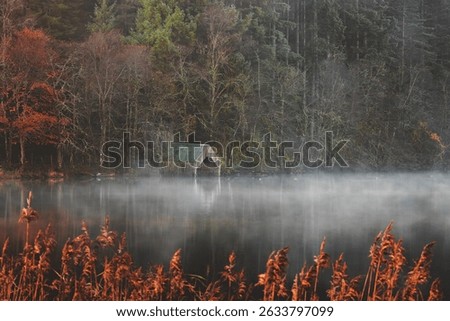 A rustic boathouse reflects in the misty, tranquil waters of Loch Ard, creating an eerie yet serene autumnal scene in Aberfoyle, Scotland.