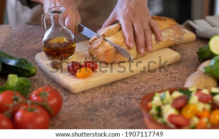 A rustic bread on the wooden cutting board and two woman's hands preparing a snack with a tomato bruschetta and spicy oil. Assortment of fresh vegetables on the table