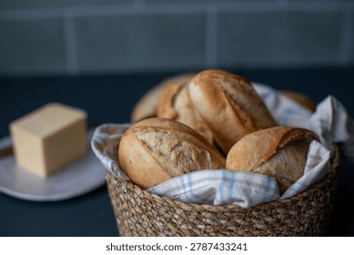 Rustic breakfast setting featuring crisp artisan French buns in a woven bread basket on a dark kitchen countertop.