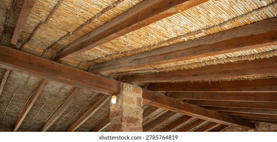 A rustic ceiling in a traditional spanish structure. Natural reeds and dark wooden beams create a warm atmosphere. A stone pillar supports the design.
