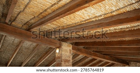 A rustic ceiling in a traditional spanish structure. Natural reeds and dark wooden beams create a warm atmosphere. A stone pillar supports the design.
