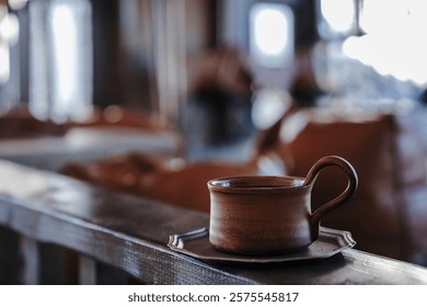 Rustic ceramic coffee cup placed on a matching saucer atop a wooden table, surrounded by a tranquil outdoor setting. The sunlight filters gently through lush green trees, creating a bokeh effect.