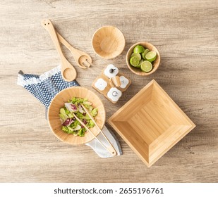 Rustic chic inspired top view of natural bamboo salad bowls, lime slices, wooden utensils, ceramic condiment jars and cloth napkins on oak surface, evoking freshness and casual rustic dining charm.