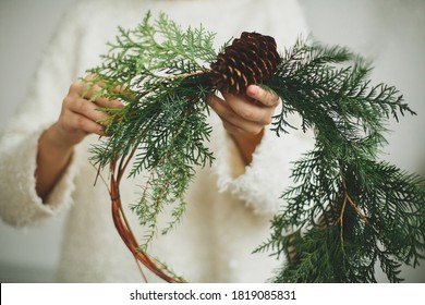 Rustic christmas wreath in female hands. Florist holding modern natural wreath with pine cone and cedar branches on white background. Seasonal holiday workshop or making wreath at home