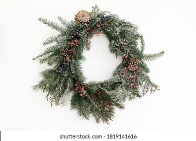 Rustic Christmas Wreath Hanging On White Wall Indoors, Festive Modern Decoration. Creative Stylish Christmas Wreath With Red Berries And Fir Branches, Isolated In White. Merry Christmas