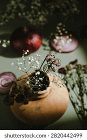 A rustic clay vase filled with delicate white baby's breath flowers and small purple blossoms sits against a muted green background. Soft natural light creates dramatic shadows while a whole red onion and sliced onion in the background add rich burgundy tones, organic textures, and depth to this elegant botanical still life composition.