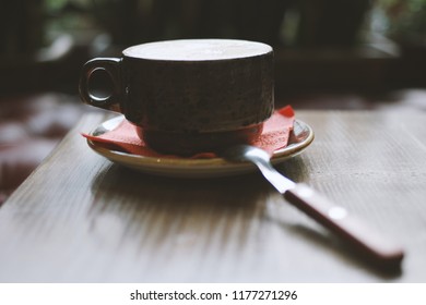 Rustic coffee set: dark stoneware cup with speckled finish sits atop textured hardwood surface, showcasing handcrafted quality and inviting warmth. Subtle red napkin peeks from beneath saucer, adding vibrant accent that contrasts with creamy palette of tans. Gleaming stainless utensil rests nearby, suggesting ritual of enjoying steaming beverage in cozy indoor environment. Soft bokeh background created by out-of-focus greenery bathes scene in diffused natural illumination, lending tranquil atmosphere ideal for contemplation. This intimate still life captures artisanal charm perfect for cafe promotions, lifestyle marketing, or culinary content creation. Close-up perspective emphasizes tactile grain and sophisticated design details, making image suitable for menu concepts and social promotions. Crafted composition evokes sense of luxury and relaxed indulgence, targeting enthusiasts of fine beverages and tasteful interior aesthetics.