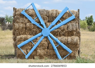 A rustic composition of stacked hay bales decorated with a large blue wooden wagon wheel in the middle of an open field. A striking rural scene under the summer sky.