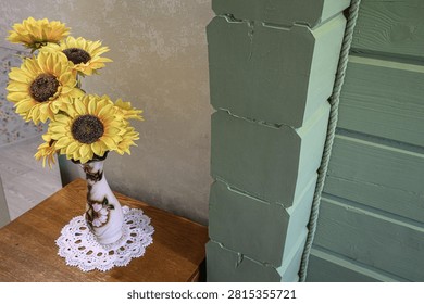 rustic corner features bright artificial sunflowers in a floral-patterned vase on a lace doily atop a wooden table, set against muted green paneled walls and a textured rope detail.