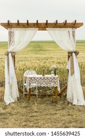 Rustic Country Chic Vintage Wooden Wedding Arbor Arch With White Curtains In An Outdoor Wedding Aisle With A Field Of Wheat In The Background