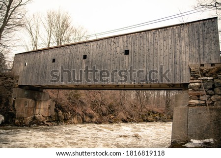 The rustic covered bridge is a timber-truss structure over the rapid river during a flood on a gloomy grey day in old sepia colors