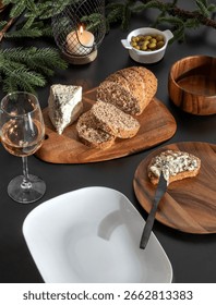 Rustic December dinner setup with whole grain bread, blue cheese, olives, rosé wine glass, fir branches, wood bowls and candlelight on dark table, evoking elegance, warmth and festive spirit.