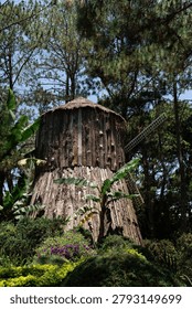 A rustic decorative windmill constructed from weathered wooden bark and topped with a thatched roof, surrounded by lush tropical vegetation. The unique structure stands amidst tall pine trees and green bushes in a scenic park under bright daylight.