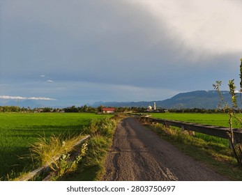 A rustic dirt path cuts through lush green agricultural fields, leading towards a distant village and majestic mountains under a dynamic, cloud-filled sky at sunset.