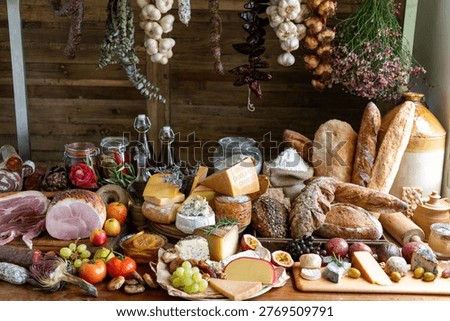 A rustic display of assorted cheeses, breads, meats, and fresh produce on a wooden table, with hanging herbs and garlic in the background.