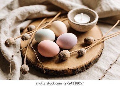 A rustic Easter arrangement featuring four pastel-colored eggs, dried poppy seed pods, and a small candle in a ceramic dish, set on a wooden slice