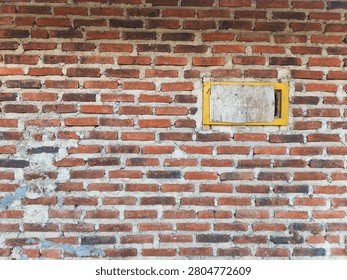 Rustic exposed red clay brick wall with natural mortar 
variation in grey and beige tones, featuring small yellow-
framed wooden shuttered window with weathered wooden panel 
closed over opening. Full frame texture of old Indonesian 
building exterior showing traditional handmade brick 
construction with visible cracks and age, concept of 
vintage architecture, rustic building materials, and 
traditional construction in Southeast Asian rural setting.