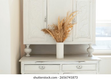 Rustic fall decor at apartment. White wooden Sideboard cupboard in home interior living room. On the buffet stands vase with bouquet dried flowers and grass. Vintage retro style. 