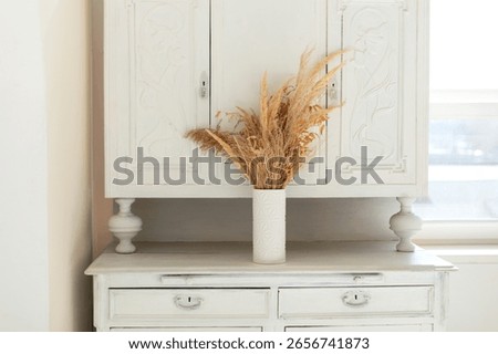 Rustic fall decor at apartment. White wooden Sideboard cupboard in home interior living room. On the buffet stands vase with bouquet dried flowers and grass. Vintage retro style. 