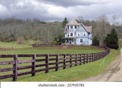 A Rustic Fence And Dirt Road By An Old Victorian Home Under An Overcast Sky In The Early Spring.  