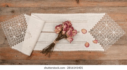 A rustic flat lay featuring a bouquet of dried pink roses placed on an elegant vintage linen table runner with intricate handmade lace inserts. Scattered rose petals and the warm wooden tabletop create a nostalgic composition celebrating traditional craftsmanship, natural beauty, and timeless home decor
