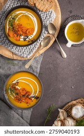 A rustic flat lay featuring two bowls of creamy carrot ginger soup topped with cream swirls, roasted carrot medallions, and black sesame seeds alongside toasted sourdough bread