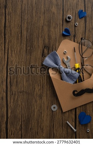 Rustic flatlay of a Father's Day gift envelope filled with tools, bow tie, glasses and paper hearts on a wooden tabletop evoking handyman charm, DIY sentiment and masculine style