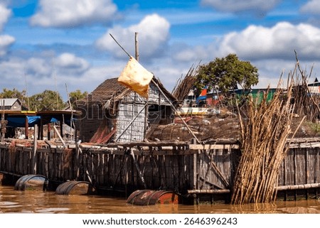 A rustic floating house sits on Tonle Sap Lake in Cambodia. The house is constructed with bamboo and wood, and surrounded by fishing nets and equipment. The barrels keep the structure afloat.