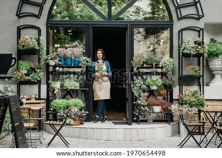 Rustic flower shop, eco cafe and business outdoor. Happy young beautiful lady waiter in apron holds bouquet of tulips for client, at front door plants studio with tables and rustic decor elements
